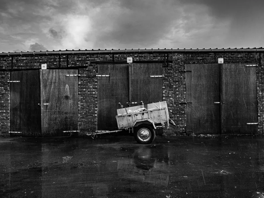 A row of old brick lock-up garages under a heavy sky, like the garage courts behind Feltham and Hanworth estates