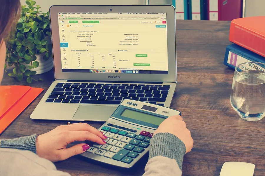 Person working out figures on a calculator in front of a laptop showing a spreadsheet