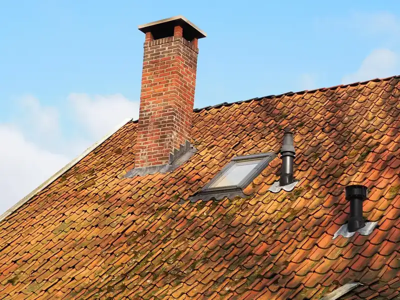 Weathered clay tile roof with a brick chimney, its metal flashing visible at the base, plus a rooflight and two vent pipes