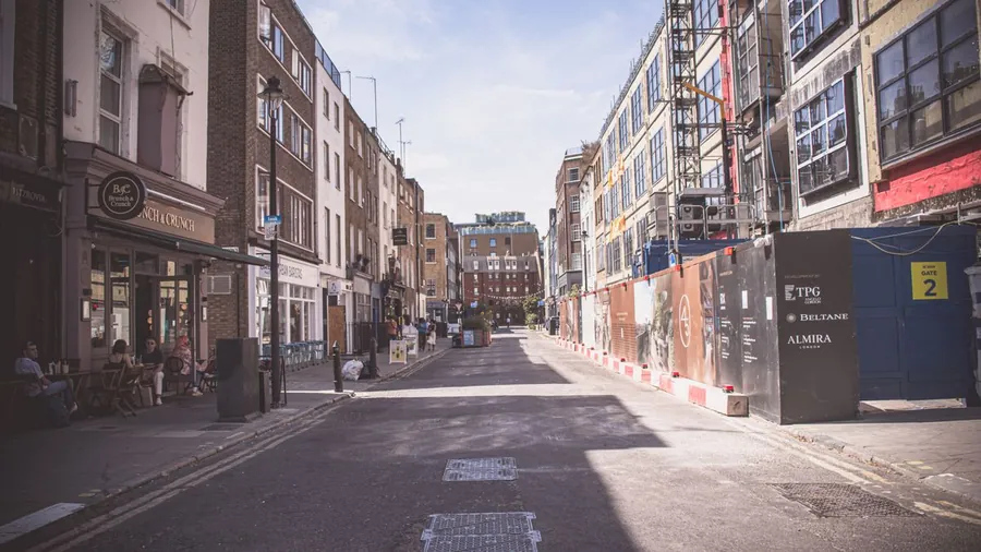 London street with shops on one side and a building wrapped in scaffolding behind site hoarding on the other