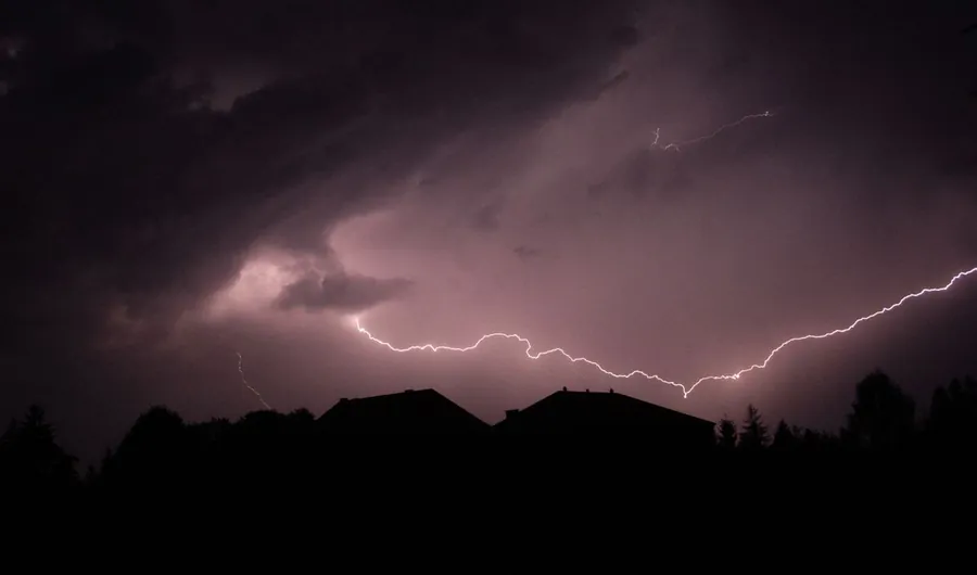 Lightning splitting a night sky above the silhouettes of suburban rooftops