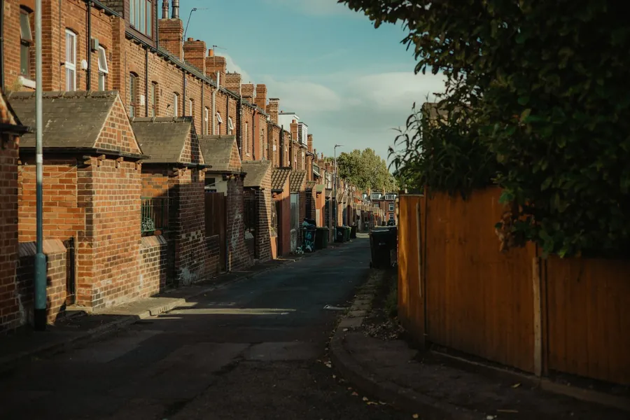 Narrow rear access lane behind a row of British brick terraced houses with wheelie bins