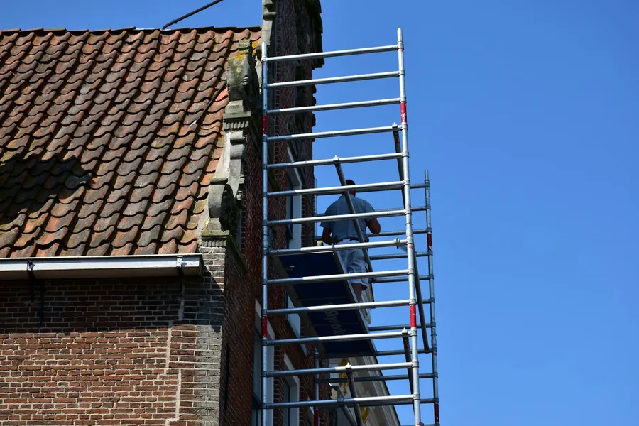 A roofer on a tower scaffold beside an old clay tile roof on a brick house, showing the access costs on a Chiswick roof job