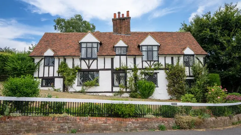 Half-timbered white cottage with steep tiled roofs behind a garden hedge